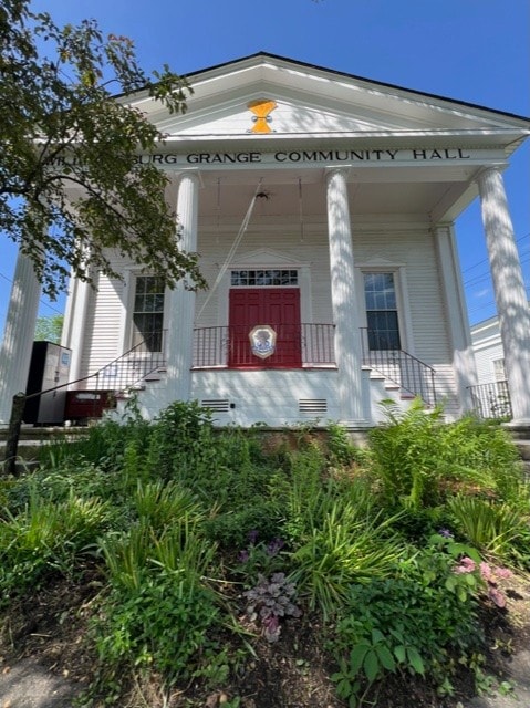 front view of a white building labeled "grange community hall" with four columns, a red door, and landscaped plants in the foreground.