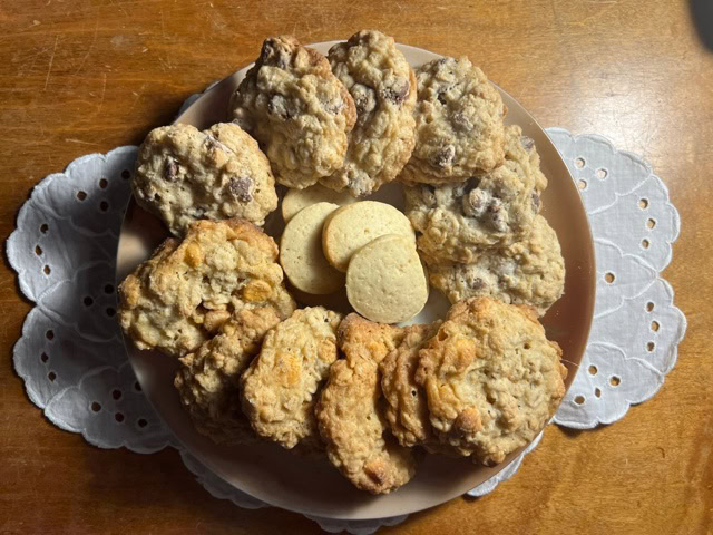 a plate of assorted cookies, including a ring of chocolate chip oatmeal cookies surrounding several plain round cookies, on a wooden table with a white doily.