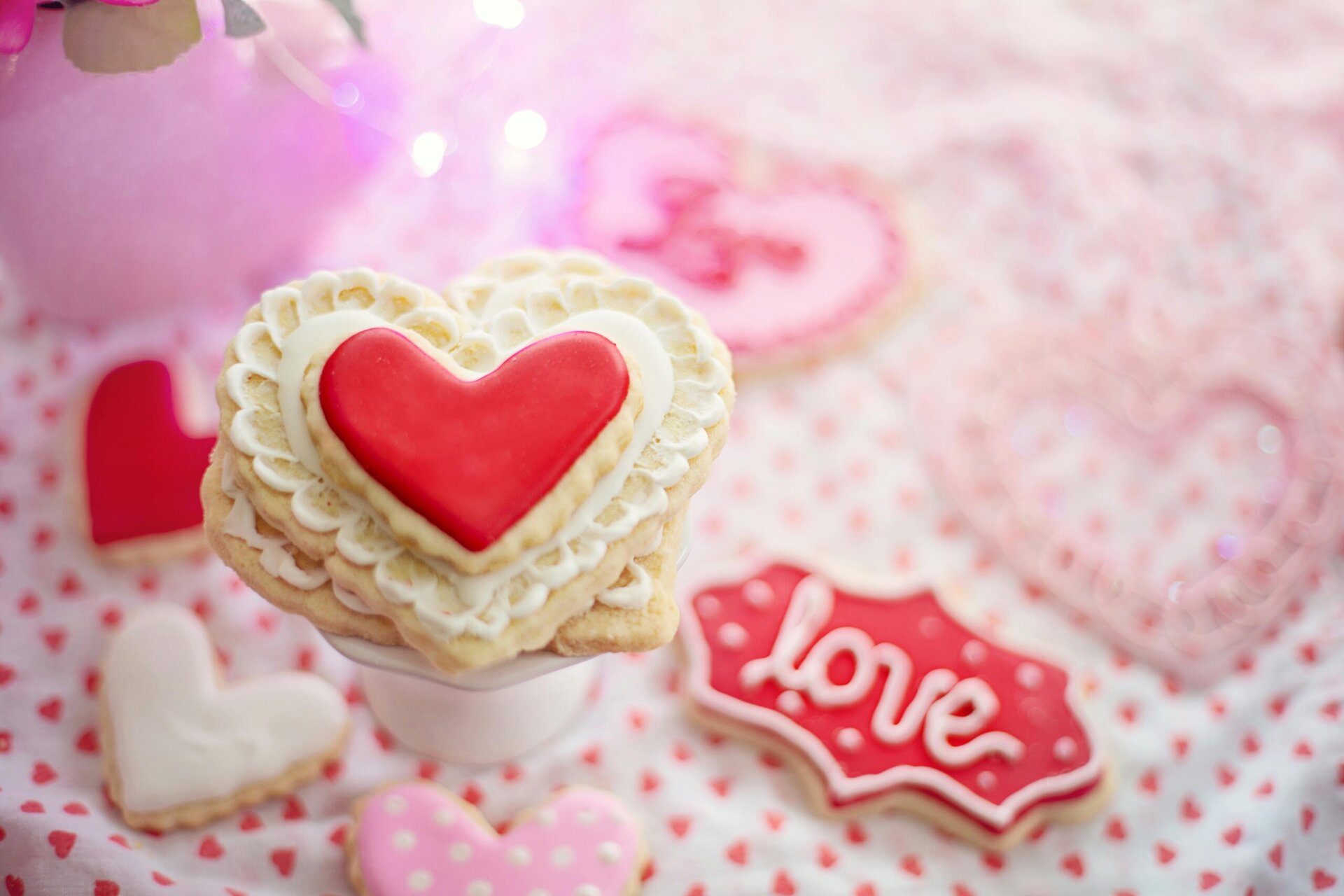 heart shaped sugar cookies with red and white icing are arranged on a pink, heart patterned tablecloth, with one cookie featuring the word "love.