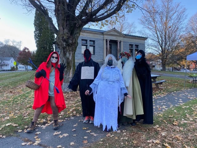 five people in halloween costumes pose outdoors in front of a historic building with bare trees and fallen leaves on the ground. one holds a sign that reads, “ghost of public records.”.