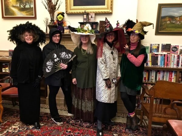 five adults in eclectic, witch themed costumes pose indoors in front of bookshelves and framed art in a library or living room setting.