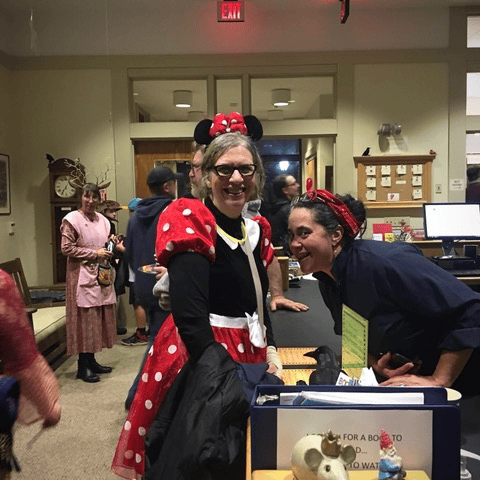 several adults are inside a library; one is dressed as minnie mouse and smiling at the camera, while others interact in the background.