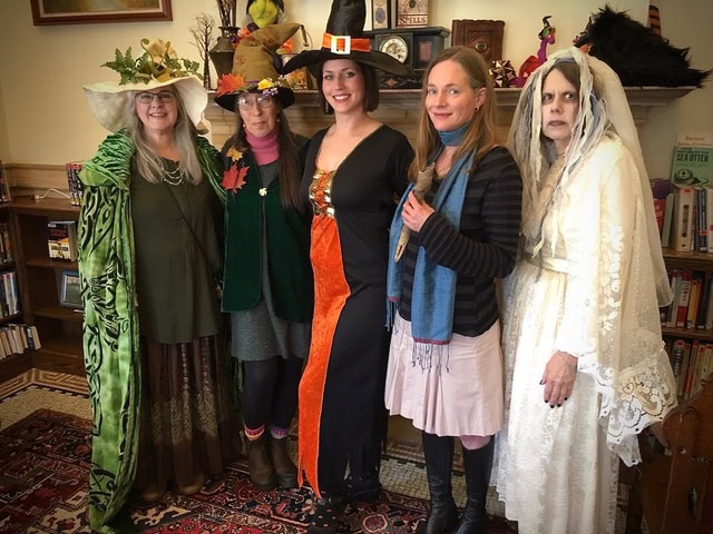 six women dressed in various halloween costumes, including witches and a ghost bride, stand together in a room with bookshelves and decorations.