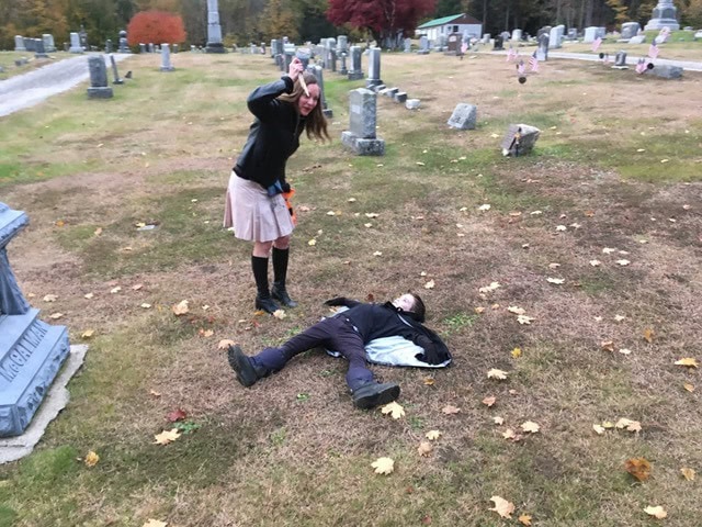 a person stands over another person lying on the ground in a cemetery. the standing person is holding a prop weapon and posing playfully. gravestones are visible in the background.