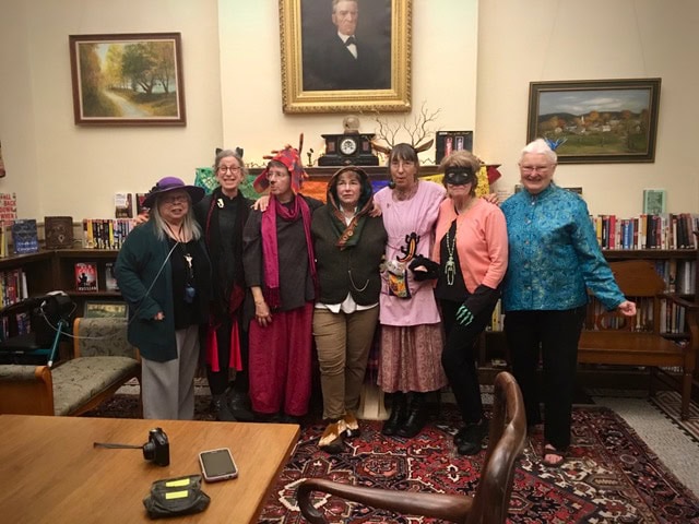 seven women in eclectic and colorful outfits pose together in a library room with bookshelves, art, and a portrait on the wall behind them.