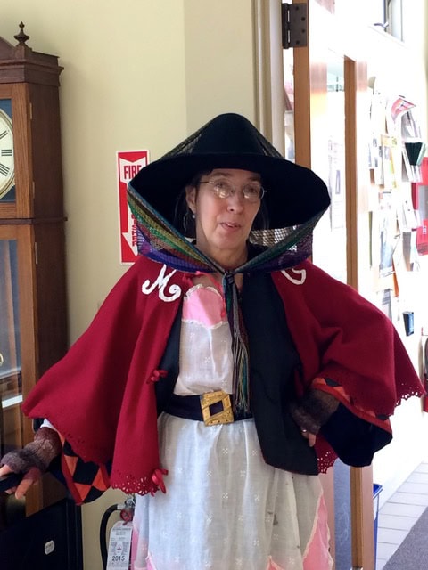 a person wearing a wide brimmed black hat, glasses, and a red cape with "mg" initials, standing indoors near a clock and a fire alarm.