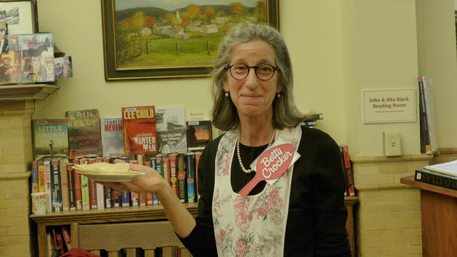 a woman wearing glasses, an apron, and a "betty crocker" ribbon stands in front of bookshelves, holding a plate with a pie or pastry, in a reading room.