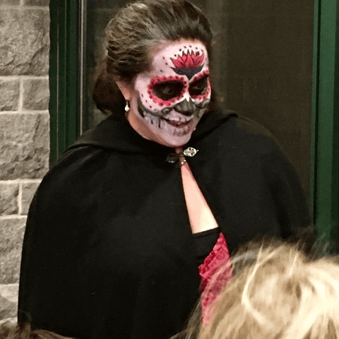 person dressed in black cape with face painted in a colorful, skull like design, resembling a day of the dead (día de los muertos) theme, standing indoors near a wall.