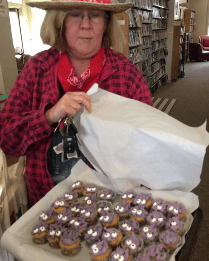 a person in a plaid shirt and red bandana holds a tray of cupcakes with purple frosting and candy eyes in a library setting.