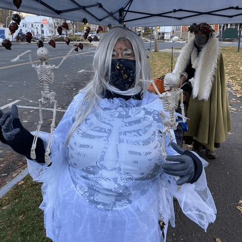 person in a skeleton costume and white wig holds plastic skeleton decorations at an outdoor event, with another person in a cloak and fur in the background under a canopy.
