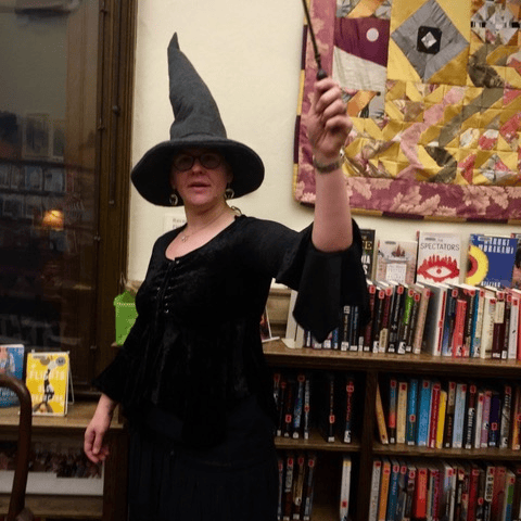 person wearing a black witch costume and hat holds a wand while standing in front of bookshelves in a library or bookstore.