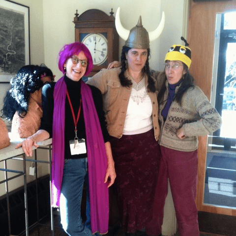 three adults pose indoors; one wears a pink wig and scarf, another a viking helmet, and the third a bee hat. a clock and a table with wigs are visible in the background.