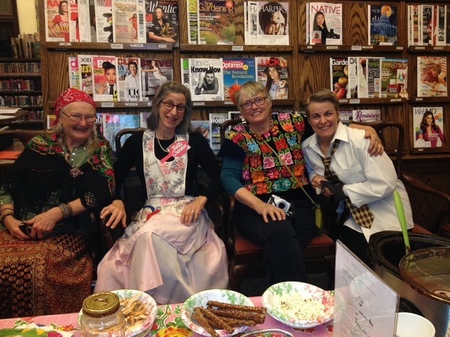 four women in colorful, festive clothing sit together and smile in front of a magazine rack at a library, with snacks and a decorated table in the foreground.