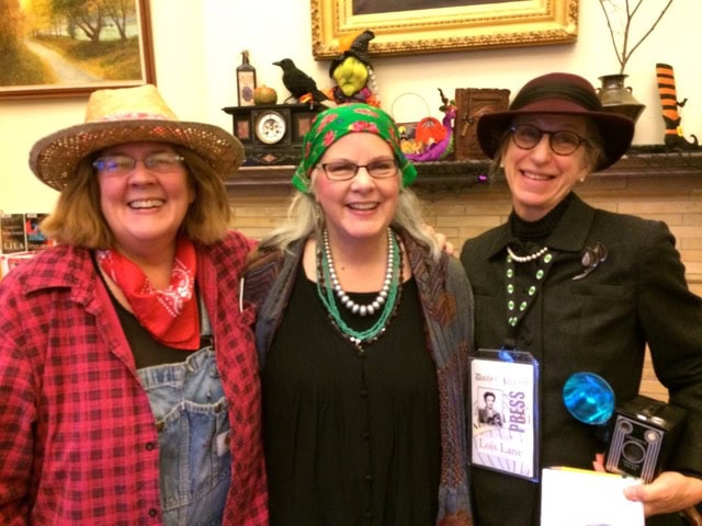 three women in costumes stand smiling together indoors, with decorative items and framed art on a mantle behind them.