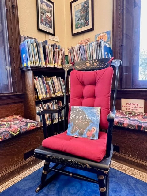 a cushioned black rocking chair with a red seat sits in a library children's section, with the book 