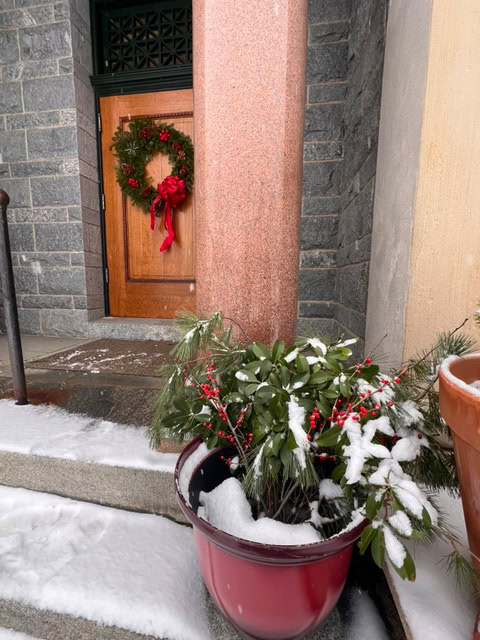 a wooden door with a holiday wreath and red bow is behind a stone column; a snow covered plant in a red pot is on the steps in front.