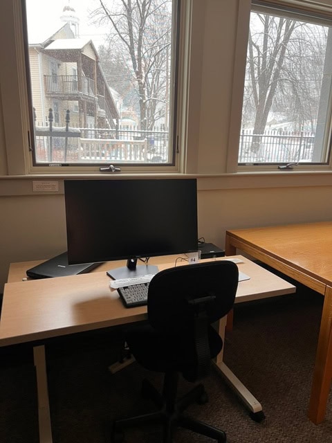 a desk with a computer monitor, keyboard, and mouse is positioned in front of windows showing trees and a snowy scene outside. an empty chair is in front of the desk.