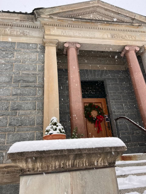 snow falls outside a stone building with columns labeled "meekins library." a wreath hangs on the door and snow covers the stairs and a potted plant.