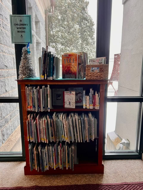 a bookshelf labeled "children's winter books" holds various children's books, a small silver christmas tree, and a window reveals a snowy scene outside.