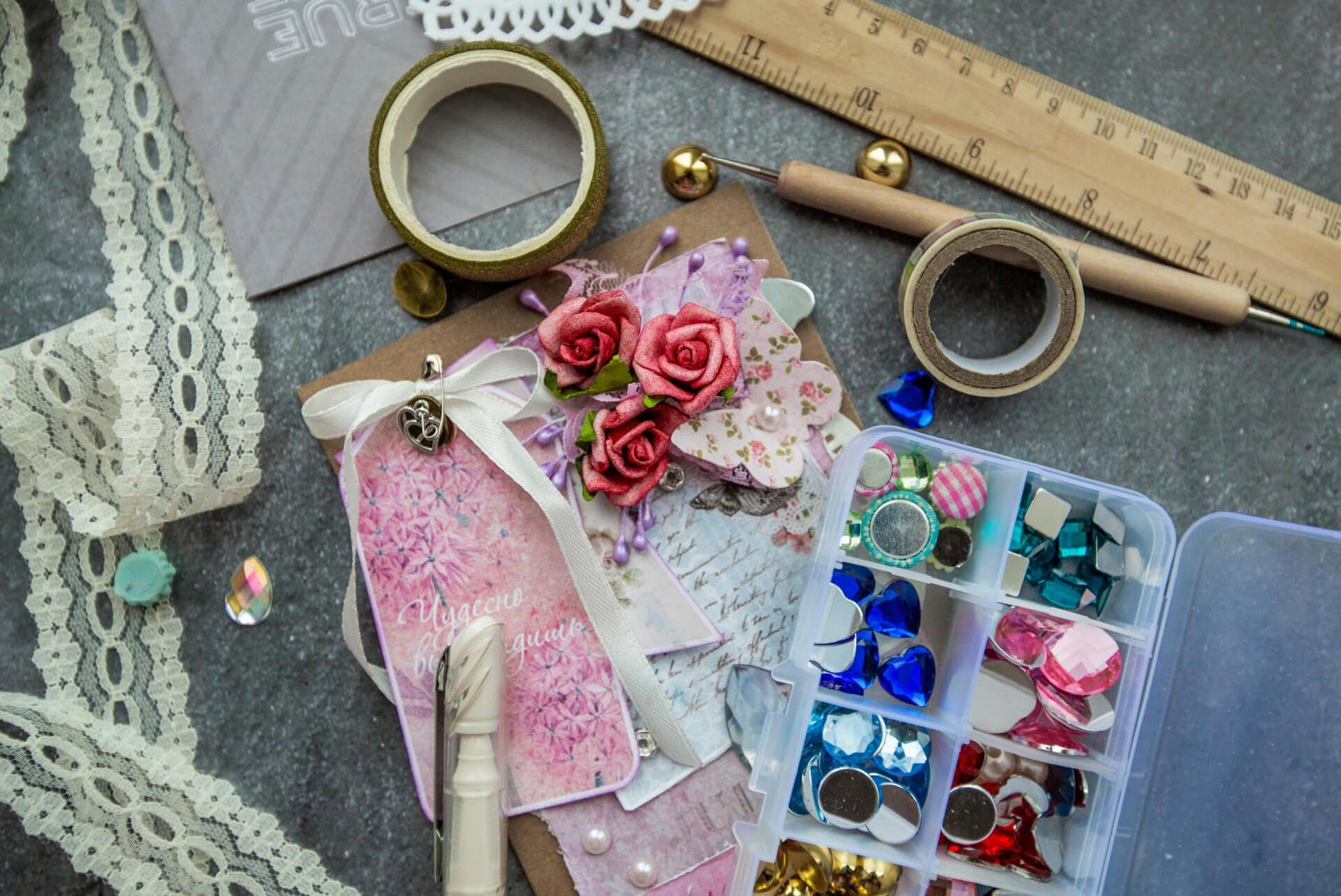 a flat lay of scrapbooking supplies, including lace, washi tape, a ruler, artificial flowers, ribbon, and a box of various colorful embellishments.