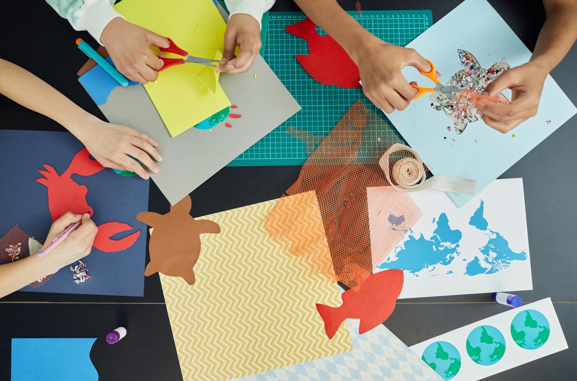 four people work together on arts and crafts, cutting animal shapes and handling paper, scissors, and various materials on a table.