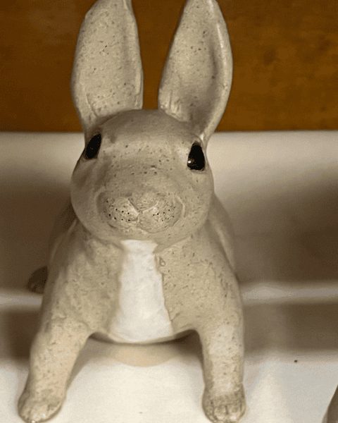 a ceramic rabbit figurine with upright ears and a white chest sits on a light colored surface against a yellow brown background.