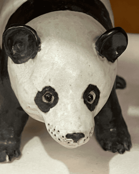close up of a ceramic or porcelain panda figurine with black and white markings, shown facing forward on a light surface.