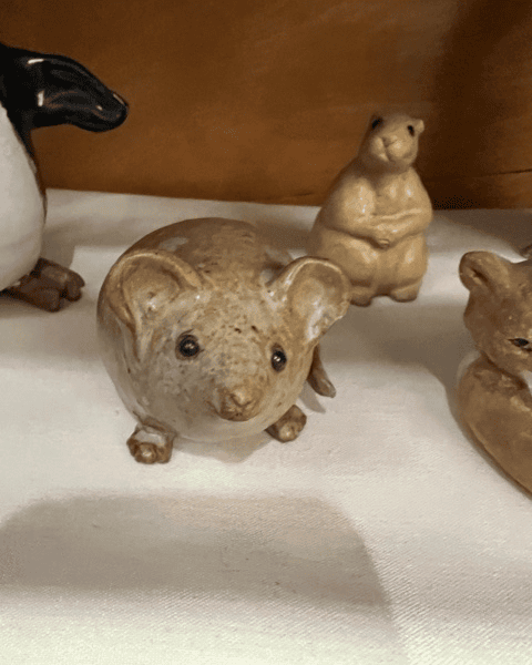 a ceramic figurine of a mouse with large ears is displayed on a light surface, with other small animal figurines in the background.