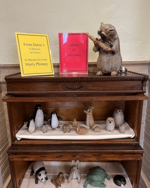 a wooden display case holds small animal figurines, with two signs on top reading “from daria’s collection of critters” and “cabinet of curiosities.” a beaver statue sits on the case.