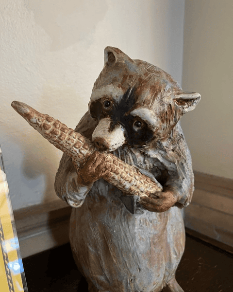 a ceramic raccoon figurine holding and biting into a corn cob, positioned on a wooden surface near a wall.