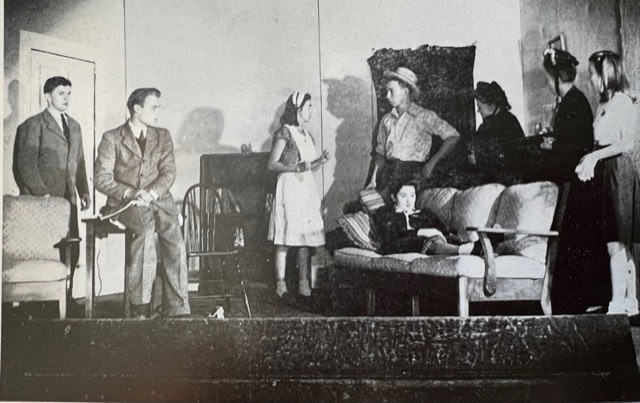 six people in vintage clothing are on stage, some standing and some sitting on a couch, appearing to perform a scene in a theatrical play set in a living room.