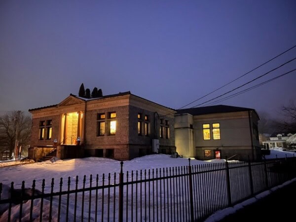 a historic stone building with glowing windows is surrounded by snow and a black metal fence at dusk.
