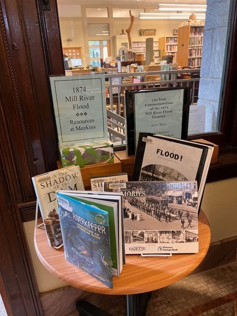 display table at a library with books and photos about the 1874 mill river flood, in front of a window with informational signs about the flood and related resources.