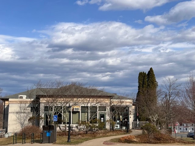 a one story building with large windows is surrounded by leafless trees and a partly cloudy sky, with a sidewalk and a blue handicap parking sign in the foreground.