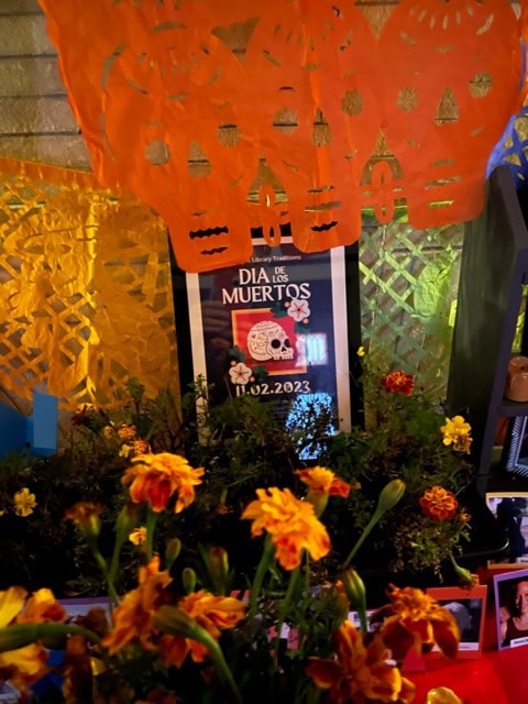 colorful marigolds, papel picado decorations, and a dia de los muertos poster with the date 11.02.2023 are displayed on an ofrenda.