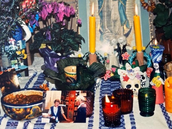 a colorful day of the dead altar with candles, flowers, food, skulls, photos, and religious imagery displayed on a striped cloth.