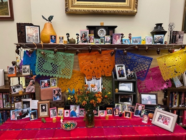 a colorful día de los muertos altar with framed photos, papel picado banners, flowers, candles, and offerings, arranged on a table and shelf against a wall with books and decorations.