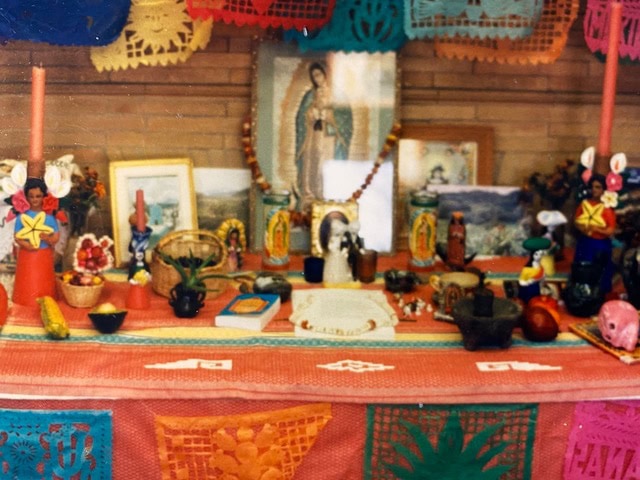 colorful day of the dead altar with candles, papel picado, religious images, small figurines, food offerings, and decorative items arranged on a striped cloth.