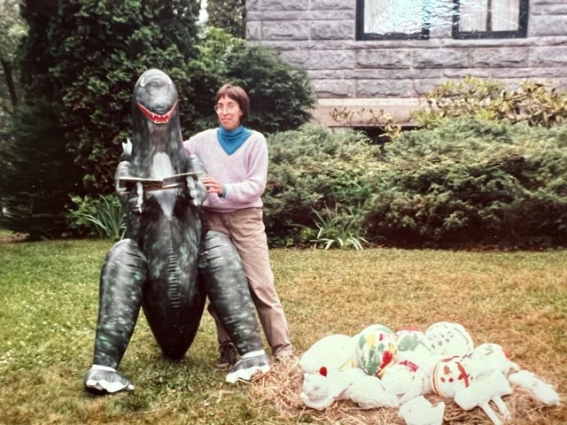 a person stands next to a large dinosaur statue holding handlebars, with several decorated round objects on straw nearby, in a garden setting.