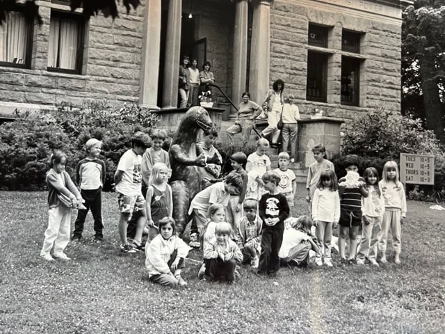 a group of children pose in front of a bear statue outside a stone building, with adults and more children standing on the entrance steps in the background.
