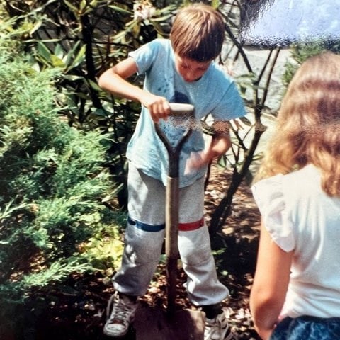 a boy stands on a shovel, pushing it into the ground, while a girl with long hair stands nearby in a garden with green foliage.