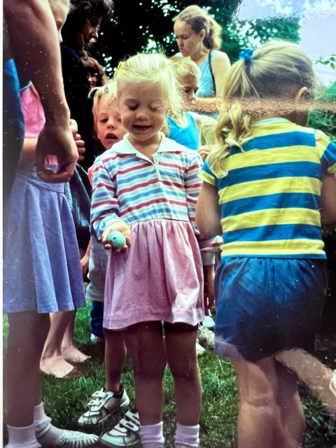 a group of children stand outdoors; a girl in a striped dress holds a blue egg while others look on. the scene appears to be during an egg hunt or outdoor gathering.