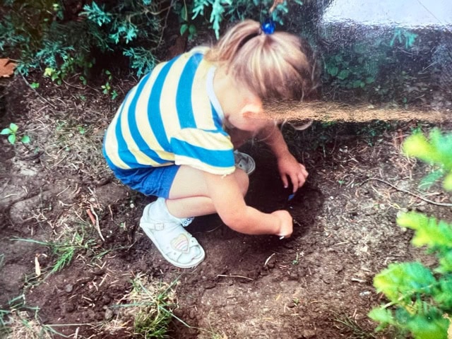 a child in a striped shirt and shorts kneels on the ground outdoors, digging in the dirt with their hands near some green bushes.