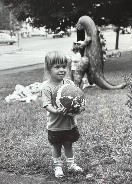 a young child stands on grass holding a worn ball, with a large dinosaur statue and other playground figures in the background.