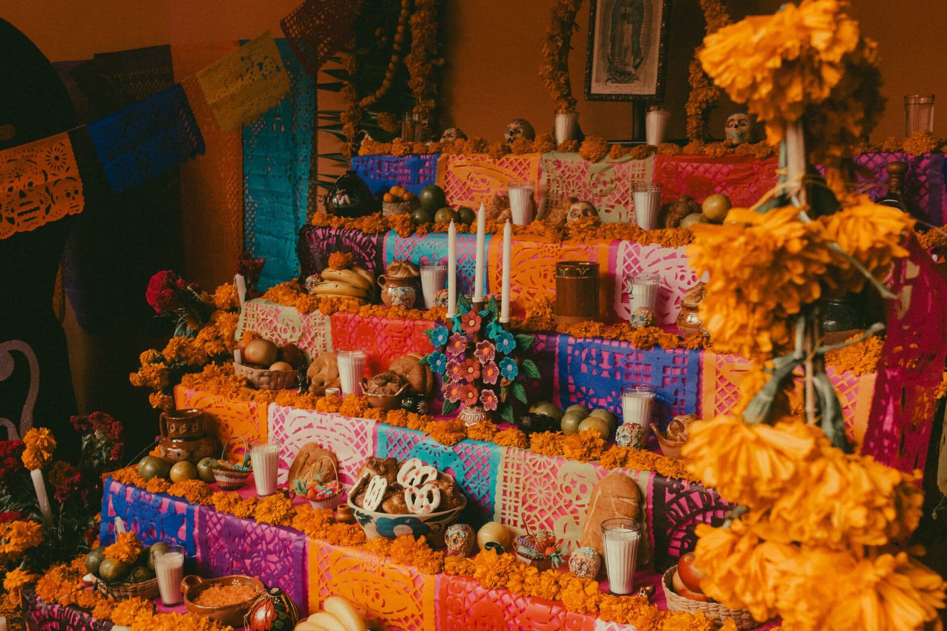a multi tiered day of the dead altar decorated with marigold flowers, candles, colorful paper banners, food, sugar skulls, and framed photos against an orange wall.
