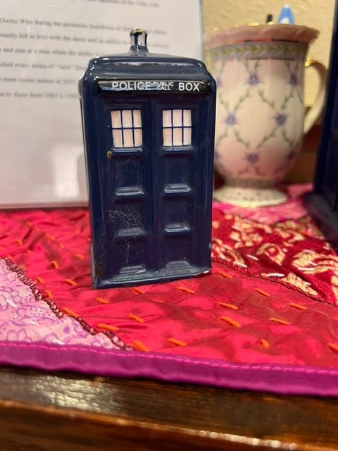a small ceramic model of a blue police public call box sits on a colorful quilted surface with a teacup and saucer in the background.