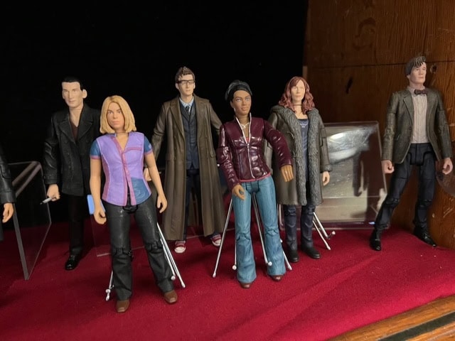 six action figures of characters from the tv series doctor who are displayed upright on a red surface, with some supported by stands.