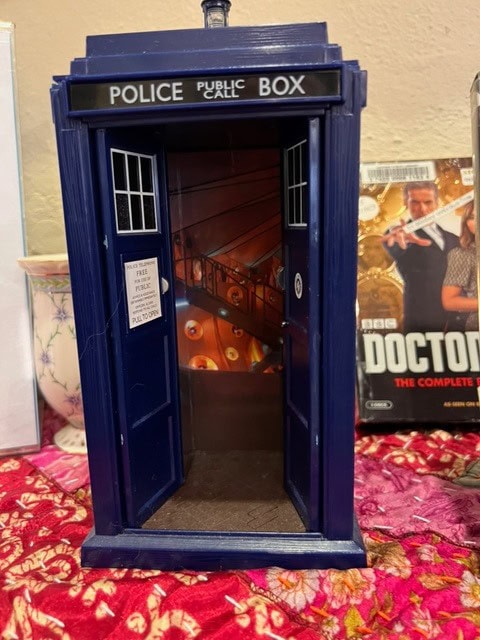 a model of the tardis from doctor who stands open on a red patterned surface, revealing an interior design. a doctor who dvd set is visible in the background.