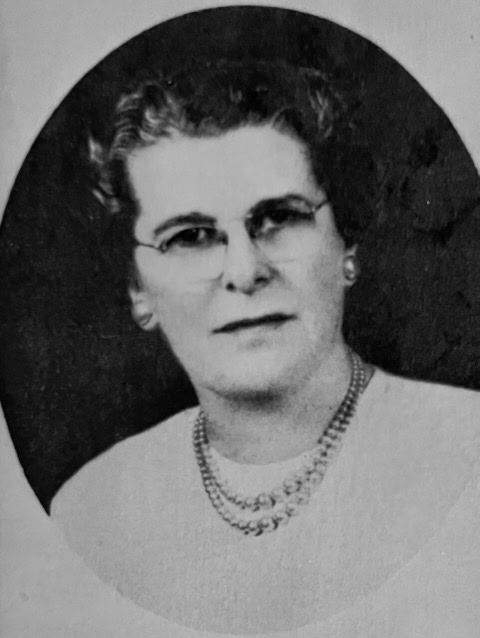 black and white portrait of an older woman with short hair, glasses, a light blouse, and a double strand pearl necklace, facing the camera with a neutral expression.