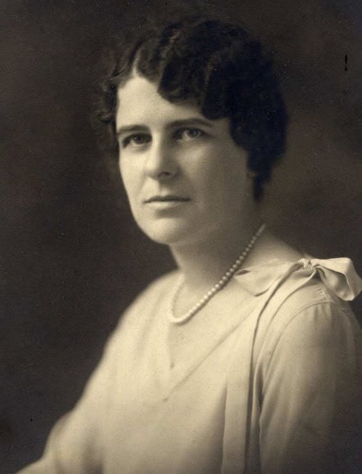 black and white portrait of a woman with short dark hair, wearing a light blouse with a bow on the shoulder and a single strand pearl necklace.